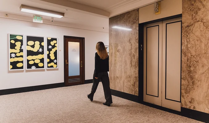 A wide-angle shot of a modern, upscale office hallway with tan carpeting and cream-colored walls. In the foreground, a closed elevator with beige doors is framed by large panels of polished brown marble. To the left, a triptych of abstract art with yellow and black patterns hangs on the wall next to a dark wood door with a frosted glass pane. A woman with long blonde hair, seen from the back, walks down the hallway toward the office entrance. The lighting is warm and professional, typical of a clinical or executive suite. - Locations in San Francisco, CA