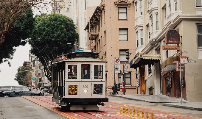 A wide-angle street view of a classic white and black San Francisco cable car traveling down a steep hill. The cable car, labeled "Powell and Market" and "Hyde and Beach," is positioned in the center on red-painted tracks. In the background, multi-story urban buildings with bay windows line the street alongside lush green trees. To the right, storefront signs for "The Last Drop Tavern" and "Mason" are visible. The scene is captured on an overcast day with soft, natural lighting. - Contact in San Francisco, CA