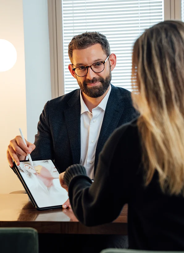 Doctor dressed in a professional dark blazer and white shirt, sits at a desk across from a patient. He is holding a digital tablet and using a stylus to point at an anatomical illustration of a face and ear, explaining a procedure during a consultation. The patient is seen from behind, showing her blonde hair and a black sweater. - Procedure in San Francisco, CA
