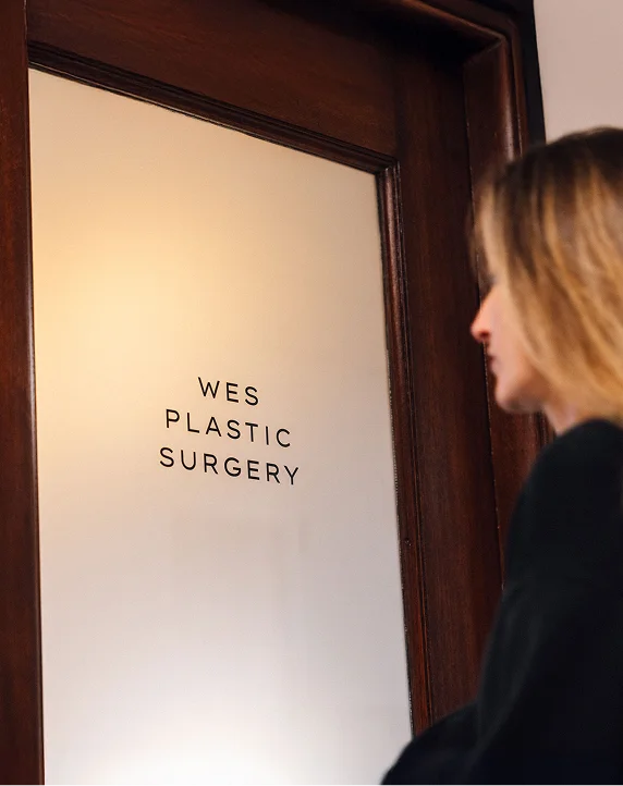 A woman stands at a dark wood door with a frosted glass pane. The text "WES PLASTIC SURGERY" is printed in clean, black capital letters on the glass. The woman is shown from the side, partially out of focus, as she looks toward the entrance. - Procedure in San Francisco, CA