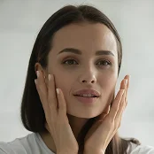 A close-up, front-facing portrait of a woman gently touching her cheeks with both hands. She has smooth skin and her hair is tucked behind her ears, appearing to examine her face in a mirror with a neutral expression. - Procedure in San Francisco, CA