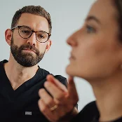 Doctor wearing black scrubs and tortoiseshell glasses, attentively examines a patient's facial profile. The patient's chin and neck are visible in the foreground, partially blurred, while the doctor remains in sharp focus against a plain white background. - procedure in San Francisco, CA