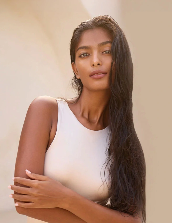 A portrait of a woman with long, dark, damp hair and a neutral expression, wearing a high-neck white top against a soft-focus beige background. - Breast augmentation with lift in San Francisco, CA