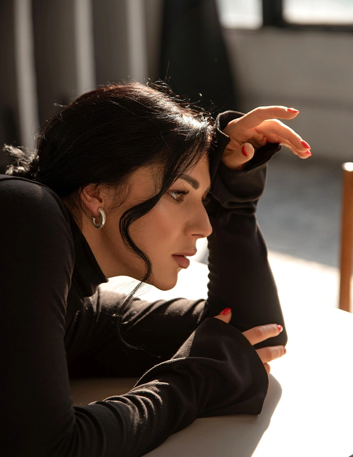 A profile shot of a woman with dark hair and silver hoop earrings, leaning her head on her arm with red-manicured nails, illuminated by soft sunlight. - Buccal fat removal in San Francisco, CA