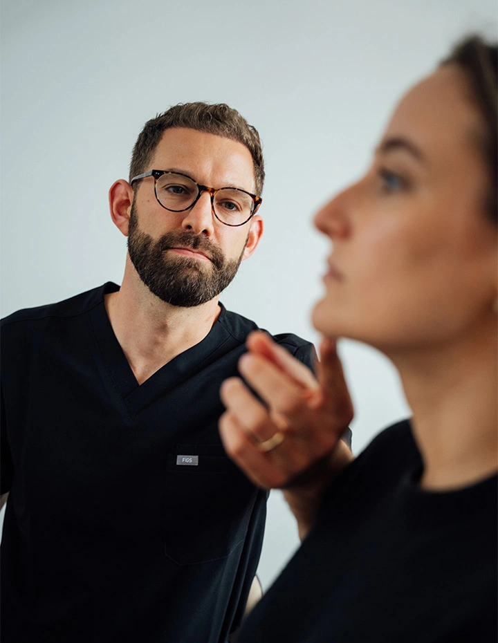 Doctor wearing black scrubs and glasses, focused on examining the chin and profile of a female patient during a clinical consultation. - Chin implants in San Francisco, CA