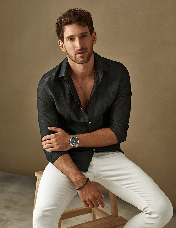 A man with short, wavy brown hair and light facial hair sitting on a wooden stool, wearing an unbuttoned dark linen shirt, white trousers, and a silver watch. - Gynecomastia in San Francisco, CA