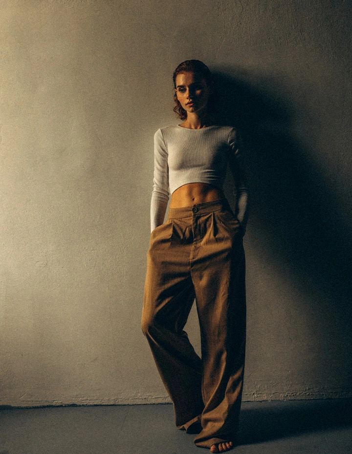 A high-fashion, moody shot of a woman with a toned midriff wearing a white long-sleeve crop top and oversized tan trousers, standing against a dimly lit, textured wall. - Liposuction in San Francisco, CA