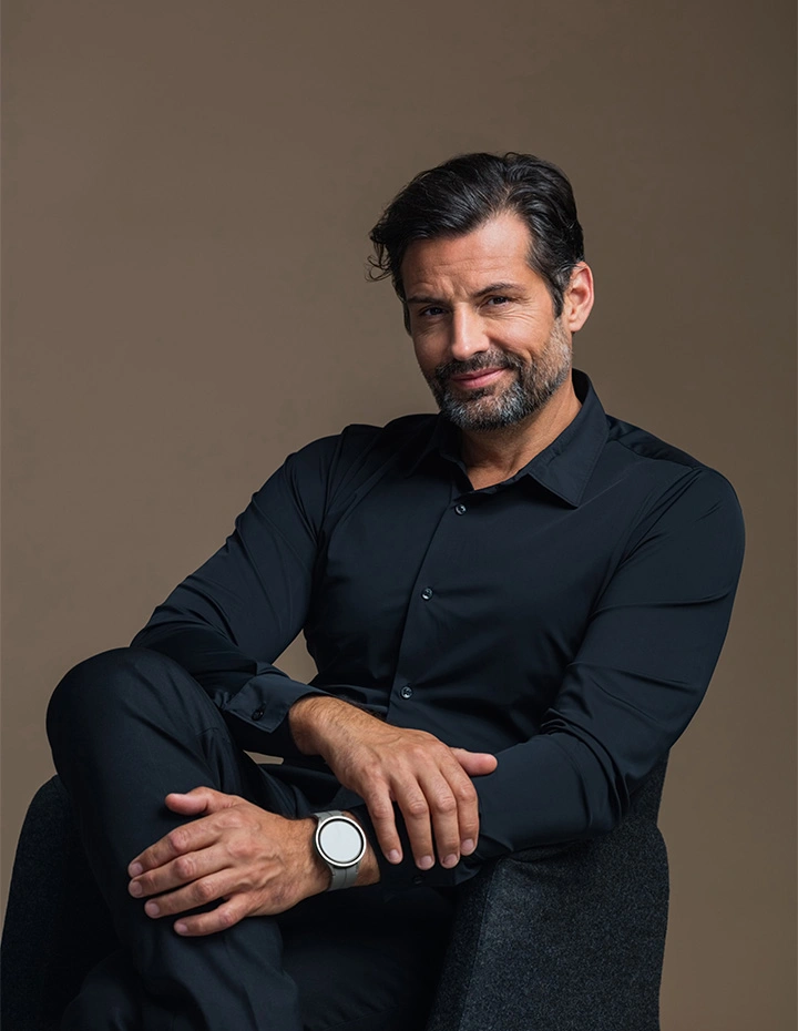 A man with dark, slightly graying hair and a groomed beard sitting in a dark shirt, resting his hands on his knee and looking directly at the camera with a subtle smile. - Male eyelid surgery in San Francisco, CA