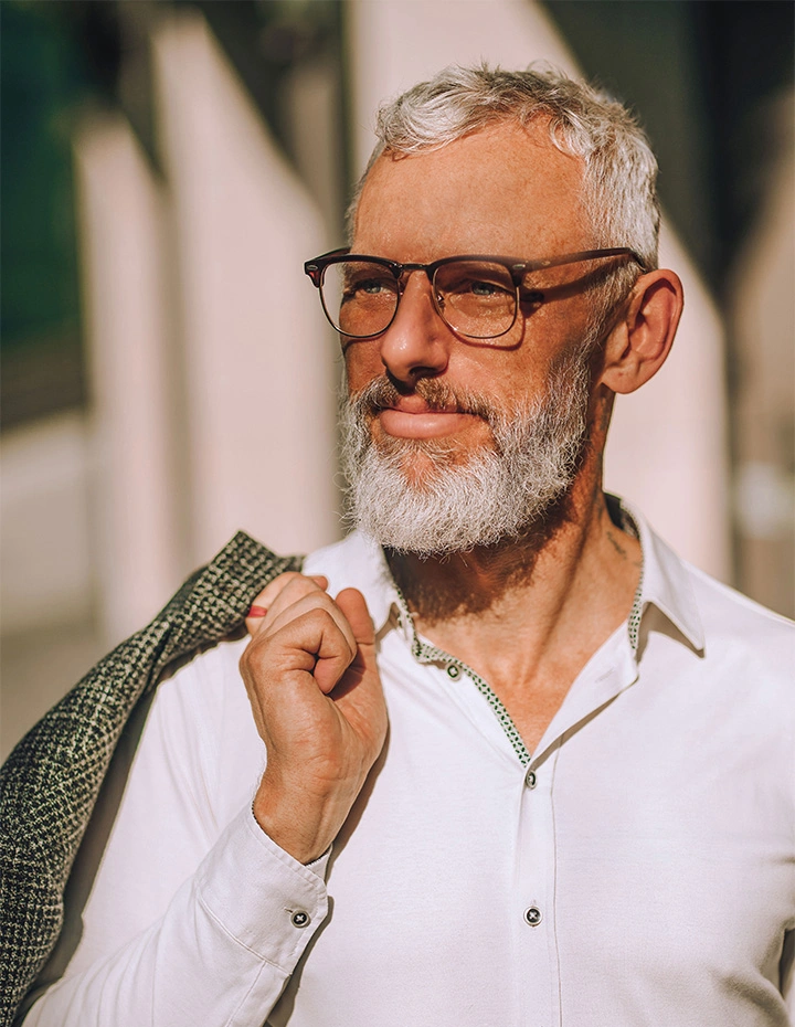 A portrait of a mature man with a well-groomed white beard and gray hair, wearing stylish glasses and a white button-down shirt with a patterned blazer draped over his shoulder. - Male necklifts in San Francisco, CA