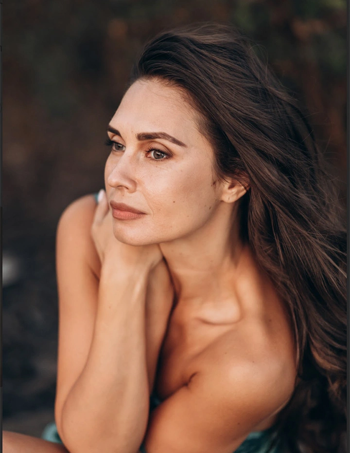 A portrait of a woman with long brown hair looking to the side, resting her chin on her hands with a soft, natural expression against a blurred background. - Brow lift in San Francisco, CA