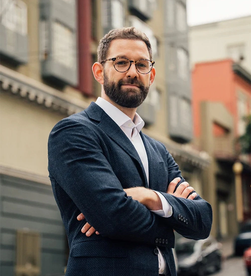 Doctor Ari Wes standing outdoors with his arms crossed. He is wearing a textured navy blue blazer over a white button-down shirt and tortoiseshell glasses. The background features a blurred urban setting with multi-story buildings under soft, natural light.