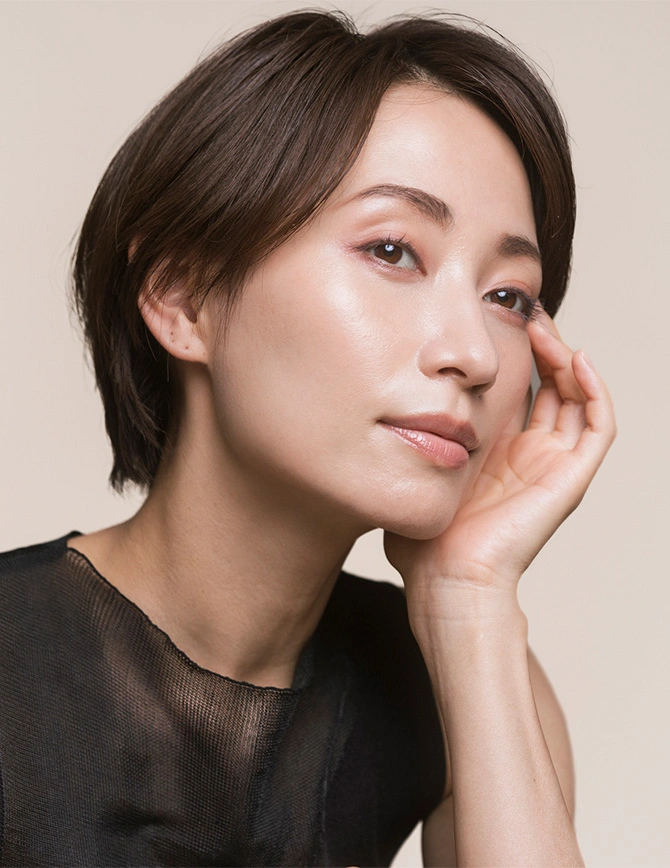 A close-up, high-angle portrait of a woman with short dark hair and glowing skin. She is resting her chin on her hand, highlighting her smooth undereye area and natural makeup look. - Eyelid surgery in San Francisco, CA