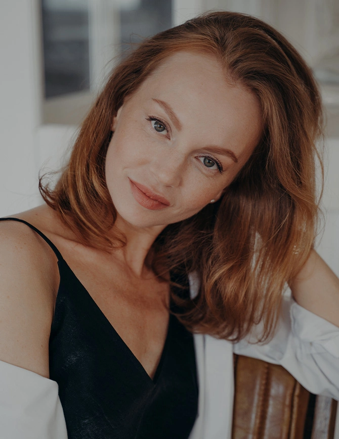 A close-up portrait of a smiling woman with vibrant red hair and light eyes. She is leaning forward slightly, wearing a black silk camisole and a white cardigan, set against a bright, airy background. - Facelift in San Francisco, CA