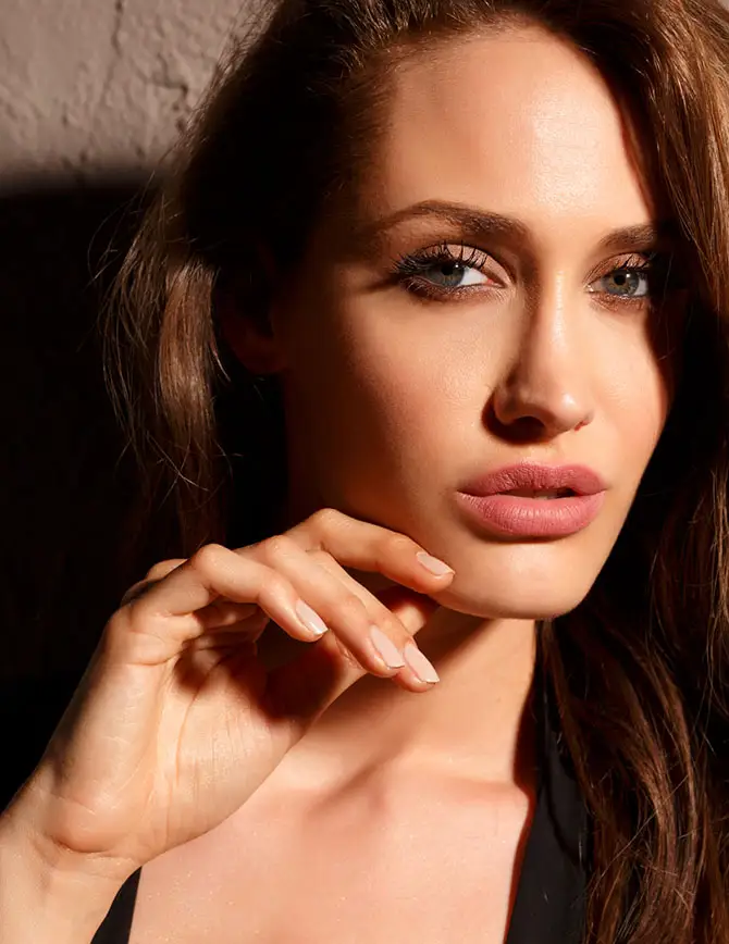 A close-up portrait of a woman with long brown hair and pink lipstick, gently resting her hand near her jawline with a focused gaze. - Facelift recovery in San Francisco, CA