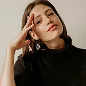 A close-up portrait of a woman with a smooth, rejuvenated complexion and subtle facial volume, wearing a black turtleneck and resting her hand gently against her forehead. - Facial fat grafting in San Francisco, CA