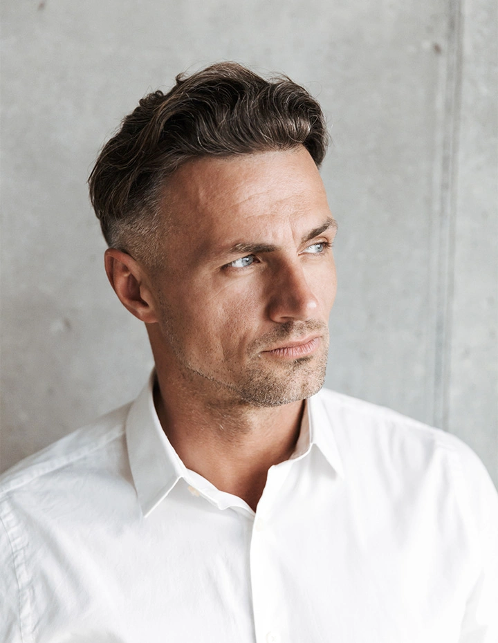 A mature man with silver hair and a groomed beard smiling thoughtfully as he looks toward a window, standing with his arms crossed in a brightly lit room. - Male facelift in San Francisco, CA