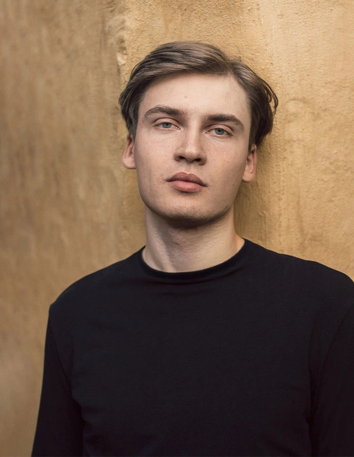 A side-profile portrait of a man with dark, curly hair and tortoise-shell glasses, dressed in a grey pinstripe blazer and black turtleneck, with dramatic lighting highlighting his facial features. - Male Rhinoplasty in San Francisco, CA